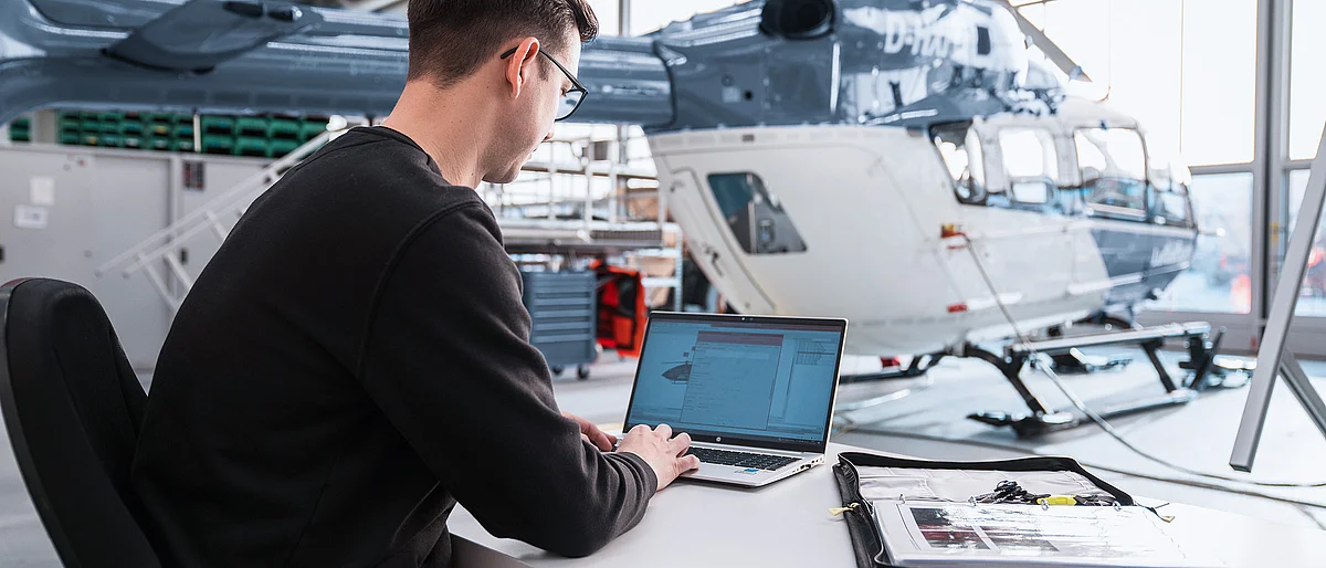 Mitarbeiter der DRF Aviation Services arbeitet an einem Laptop vor einem Hubschrauber in einem Hangar.